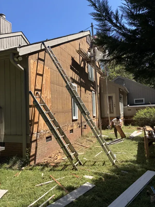 Construction worker working on house siding, two ladders leaning against wall, sunny day.