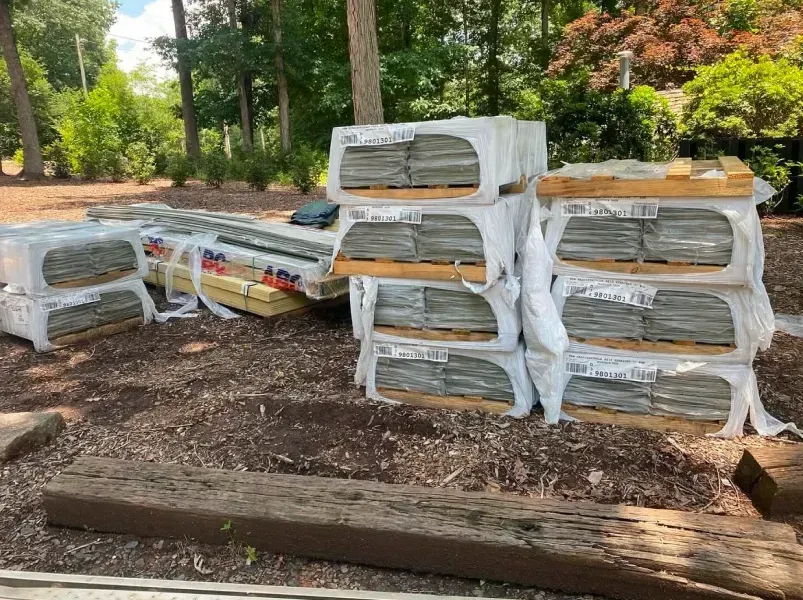 Stacks of wrapped gray pavers on pallets outdoors, surrounded by mulch and greenery.