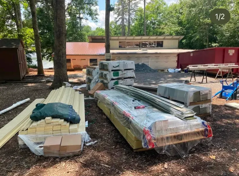 Construction site with building materials in front of a building with a red roof; trees and a dumpster in the background.