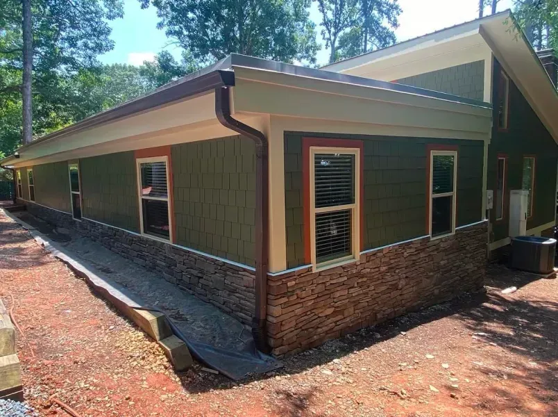 Exterior of a house with green siding, stone accents, brown trim, and a gutter system; surrounded by dirt and trees.
