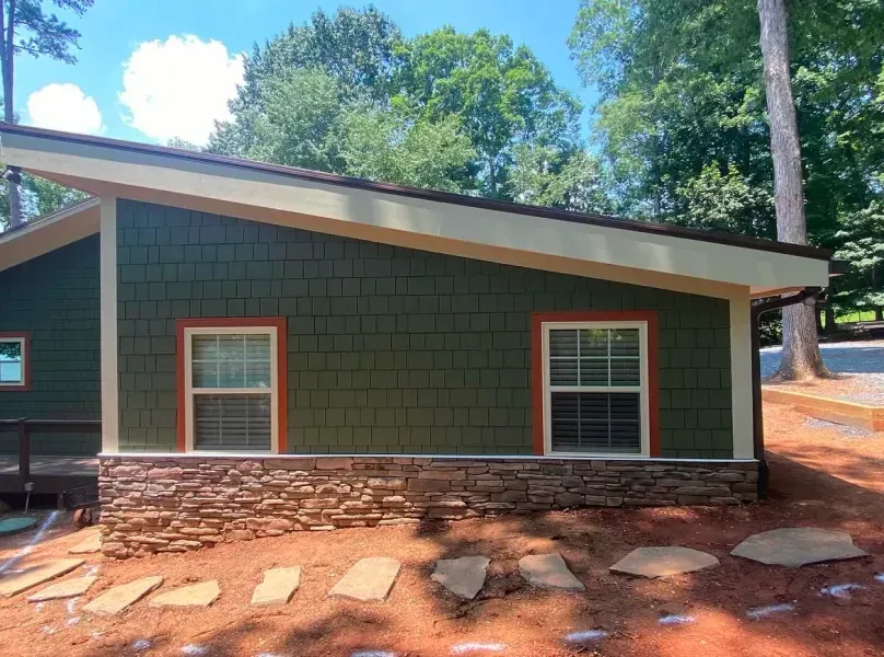 Green building with angled roof, two windows with orange trim, stone base. Outdoors, sunny.
