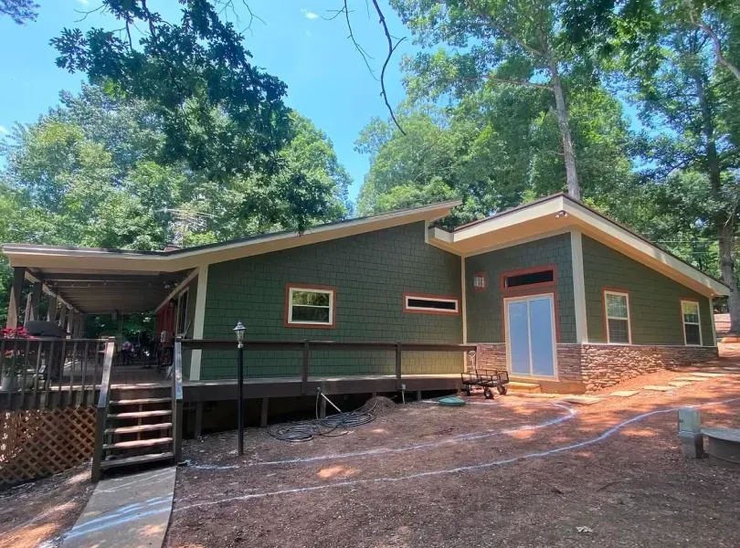 Green cabin with a deck, surrounded by trees, under a blue sky.
