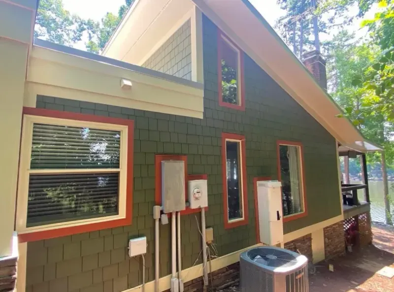 Green shingled house with orange trim, set along a lake with an air conditioning unit below.