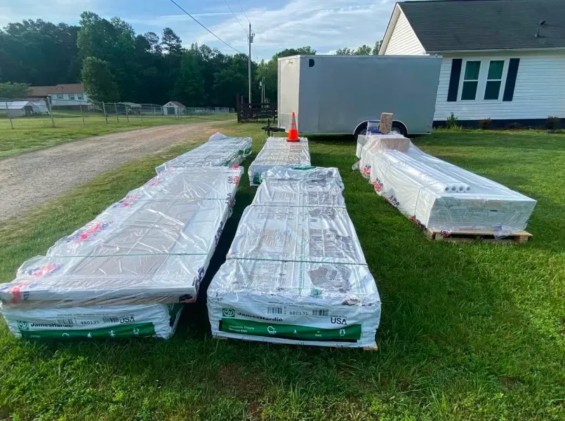 Packs of building materials wrapped in plastic sit on grass, a trailer and house in the background.