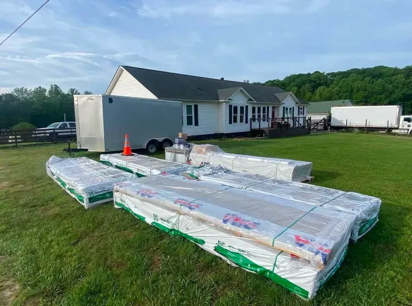Lumber packages and a trailer sit on a lawn in front of a white house on a sunny day.