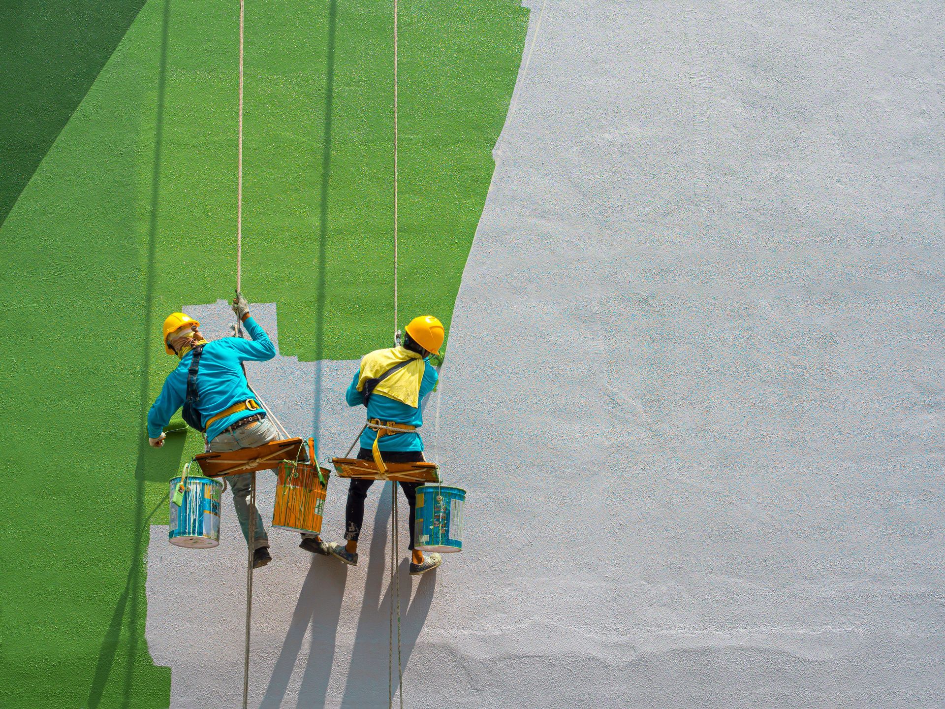 Two painters on ropes painting a wall, one green, one white.