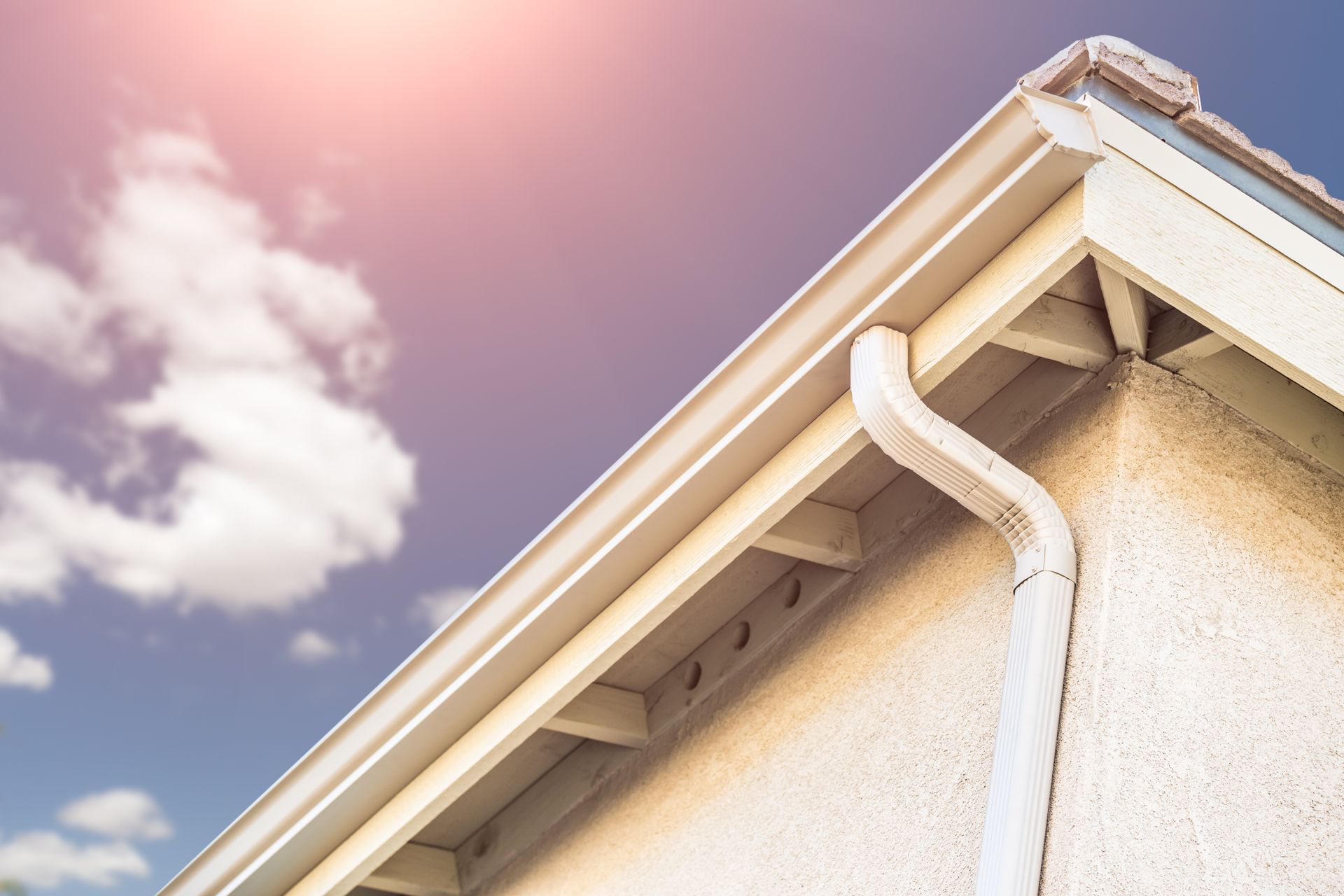 Off-white rain gutter and downspout on the corner of a light-colored stucco building, against a partly cloudy sky.