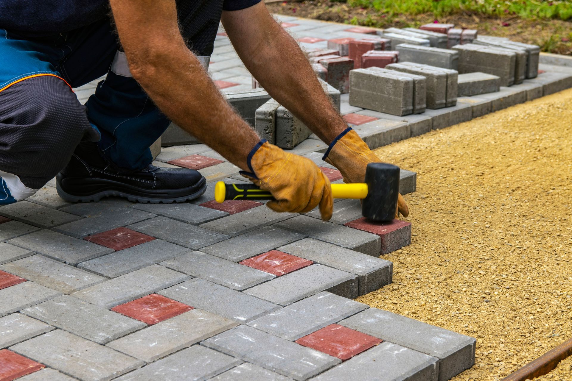 Person installing paving stones, using a mallet. Grey and red blocks, with a gravel base.