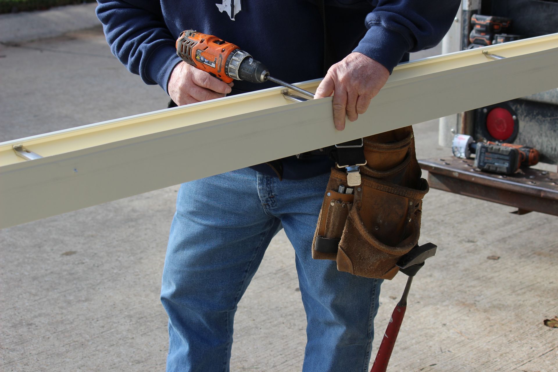 Person using a power drill to screw into a long, light-colored wooden beam, outdoors.