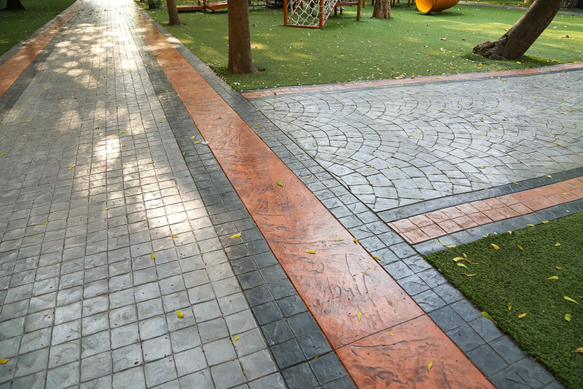 Paved walkway with textured gray blocks and an orange stripe, bordered by grass and trees.