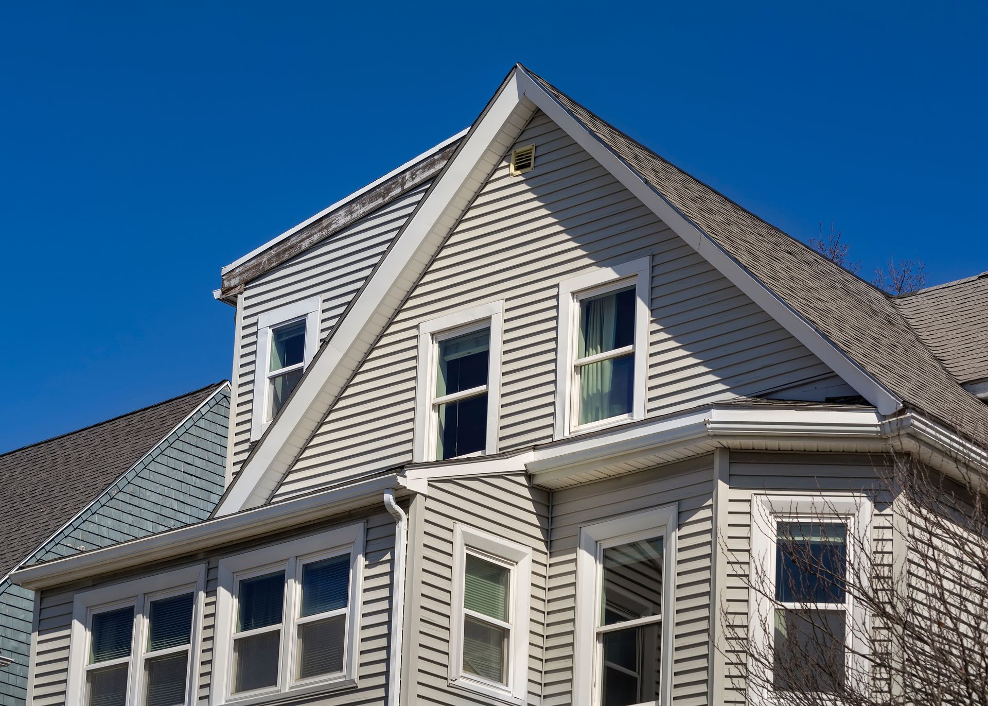 Multi-story house with gray siding, white trim, and a gabled roof against a blue sky.