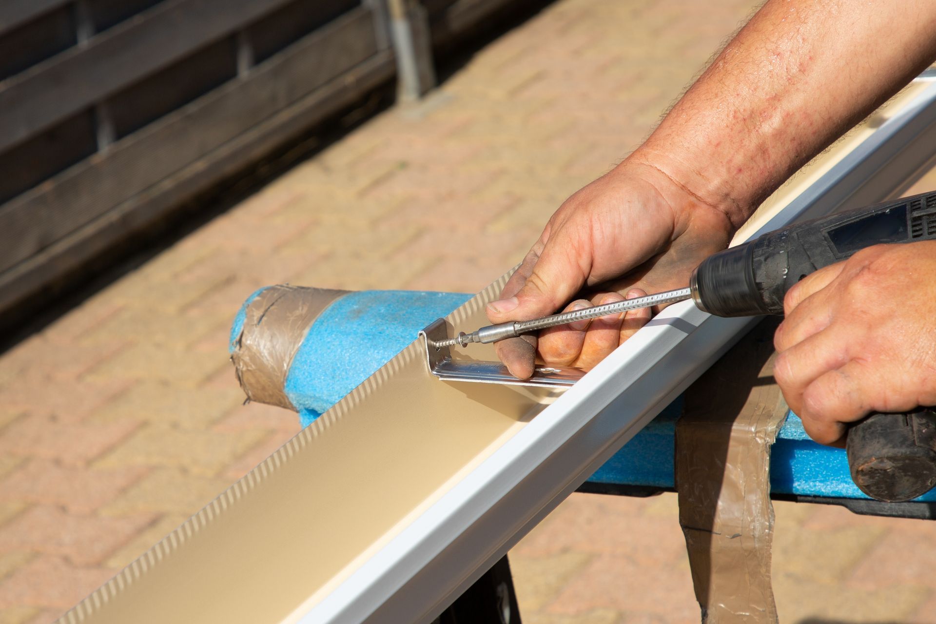 Person using a power drill to fasten a metal bracket onto a piece of tan material, outdoors.