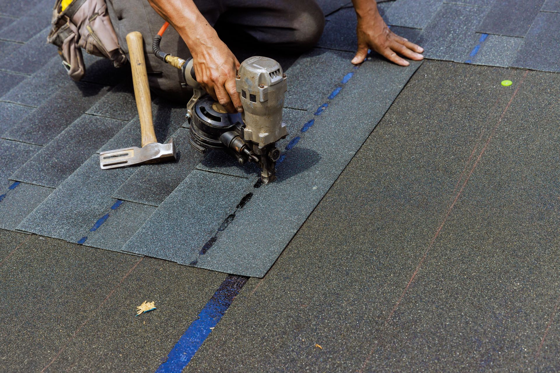 Person installing asphalt shingles on a roof, using a nail gun and hammer.