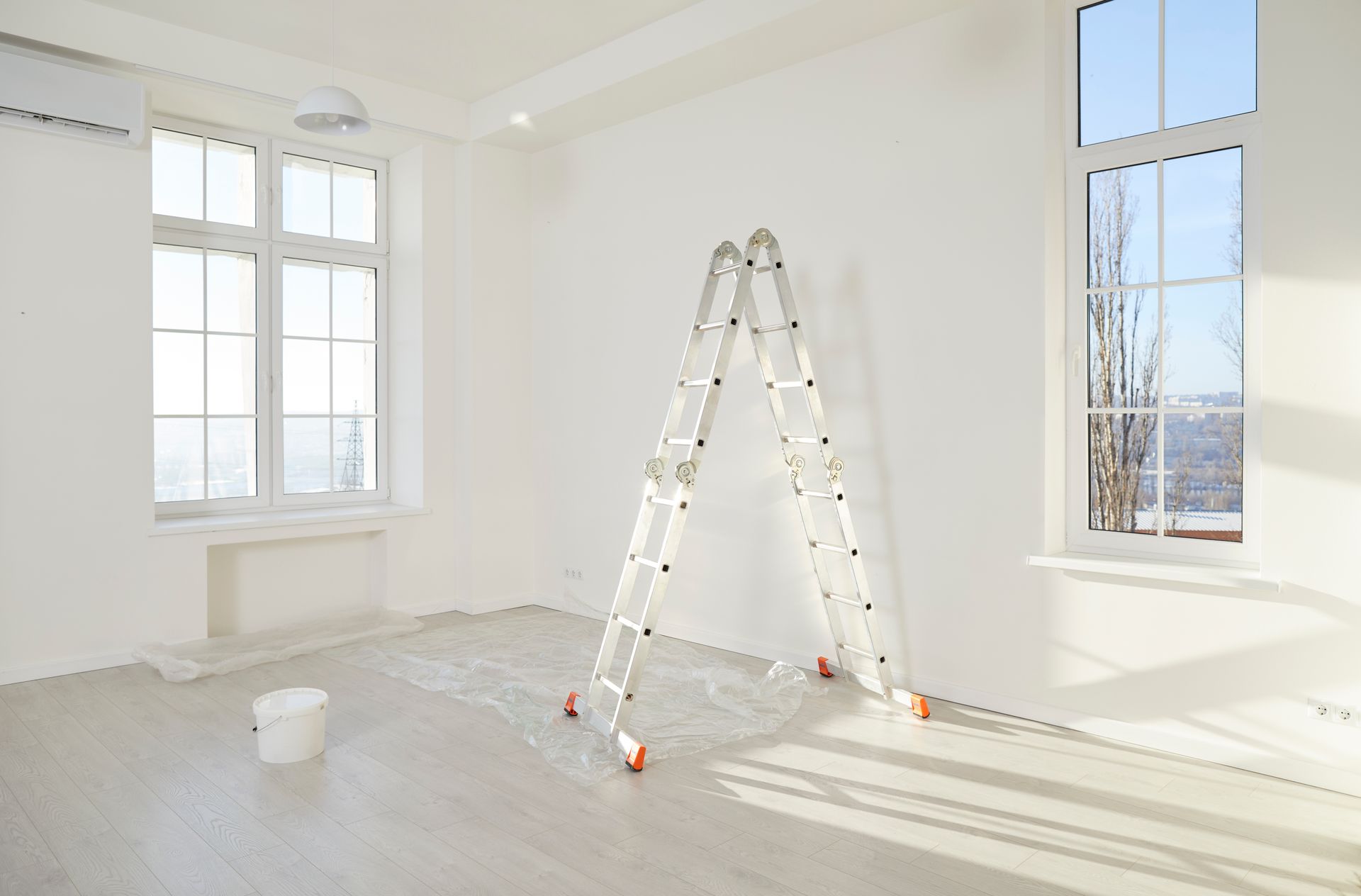 Empty room with a ladder, paint bucket, and windows; freshly painted white walls.