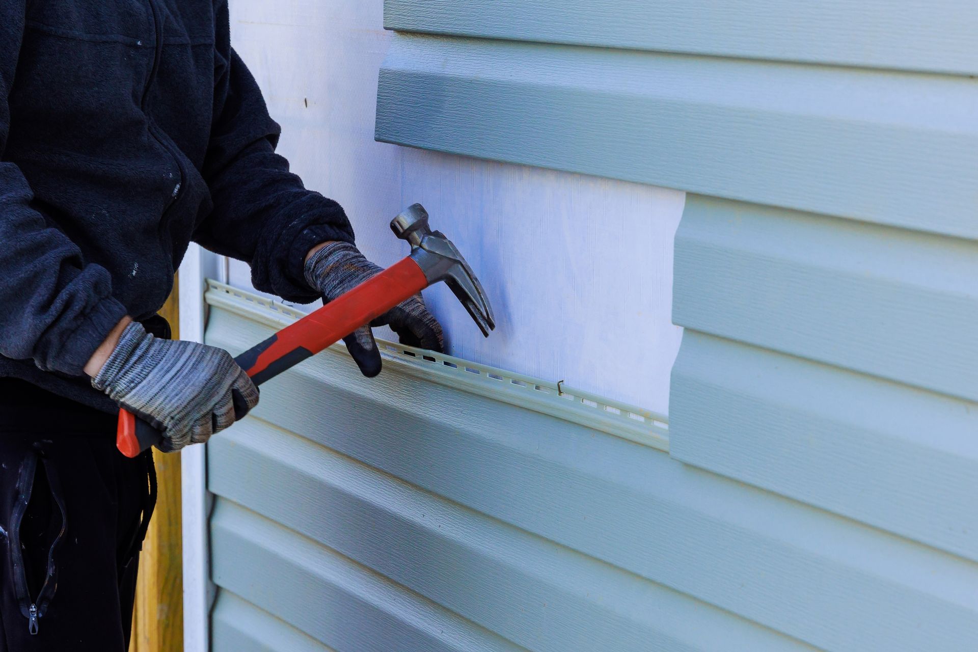 Person in gloves hammering siding on a building's exterior.