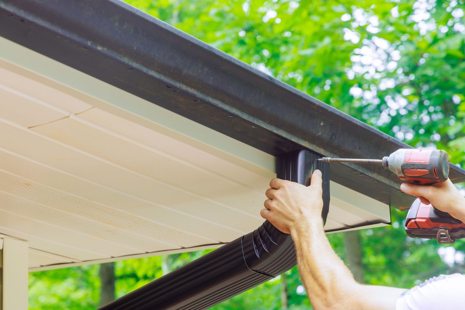 Person installing a black gutter with a power drill on a house's edge.