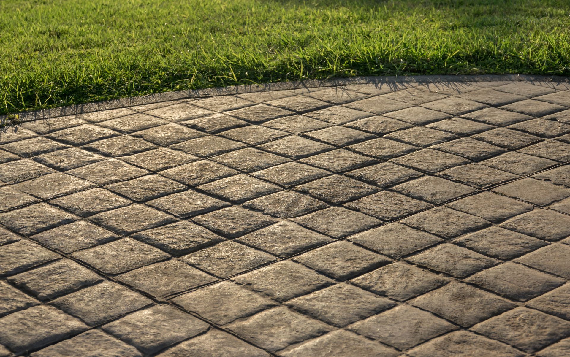 Diamond-patterned gray stone patio bordering green grass in sunlight.
