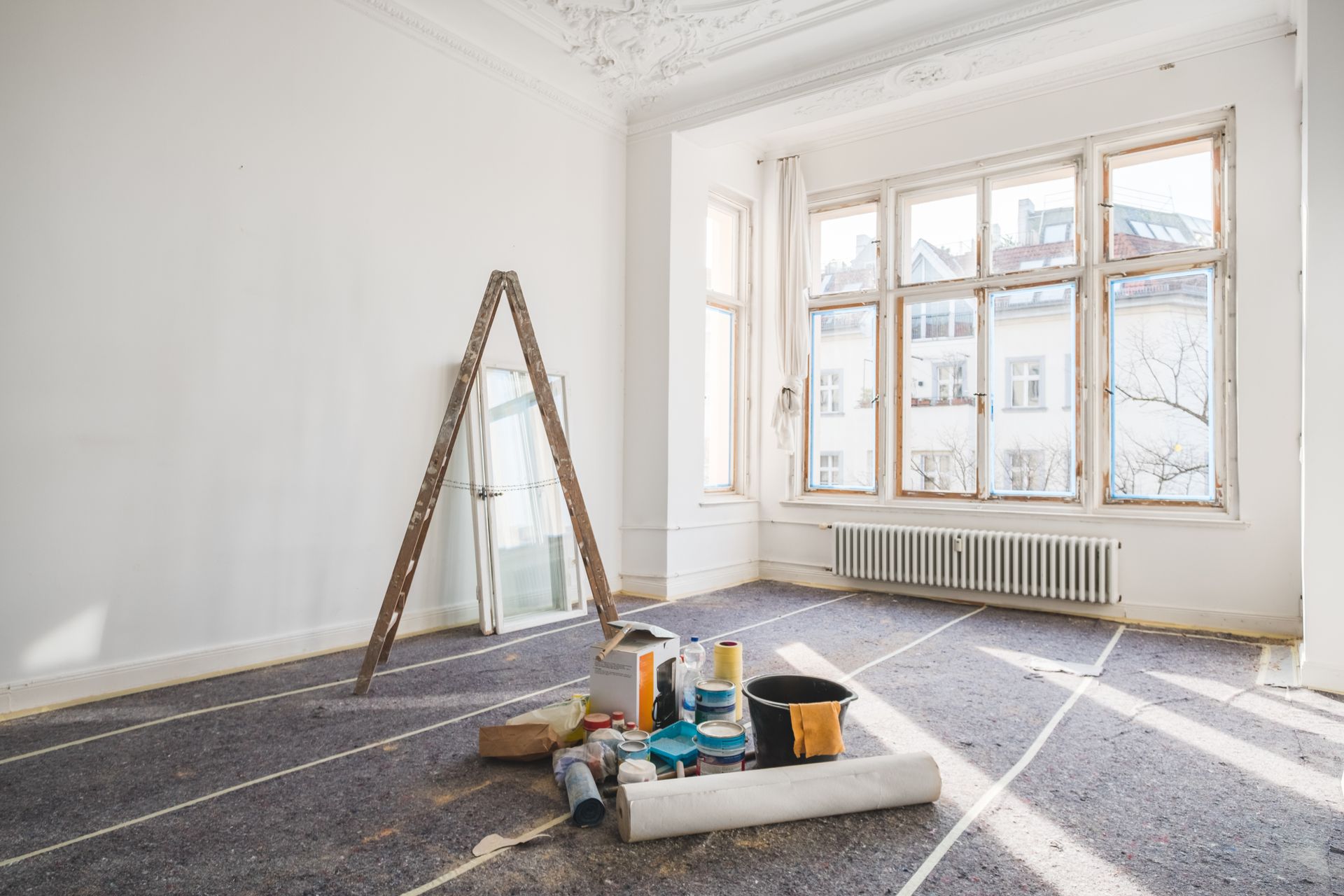 Room being renovated, with ladder, paint cans, and window; gray floor covering.
