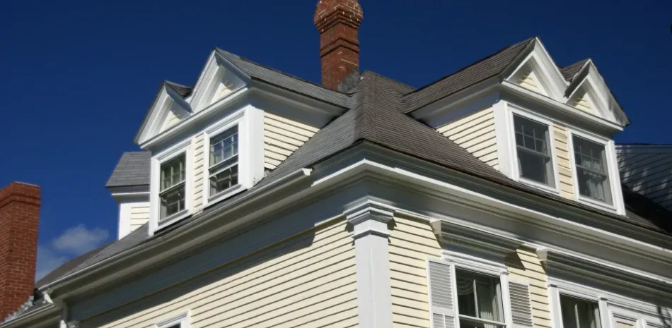 Yellow house with a dark roof and brick chimney against a blue sky.