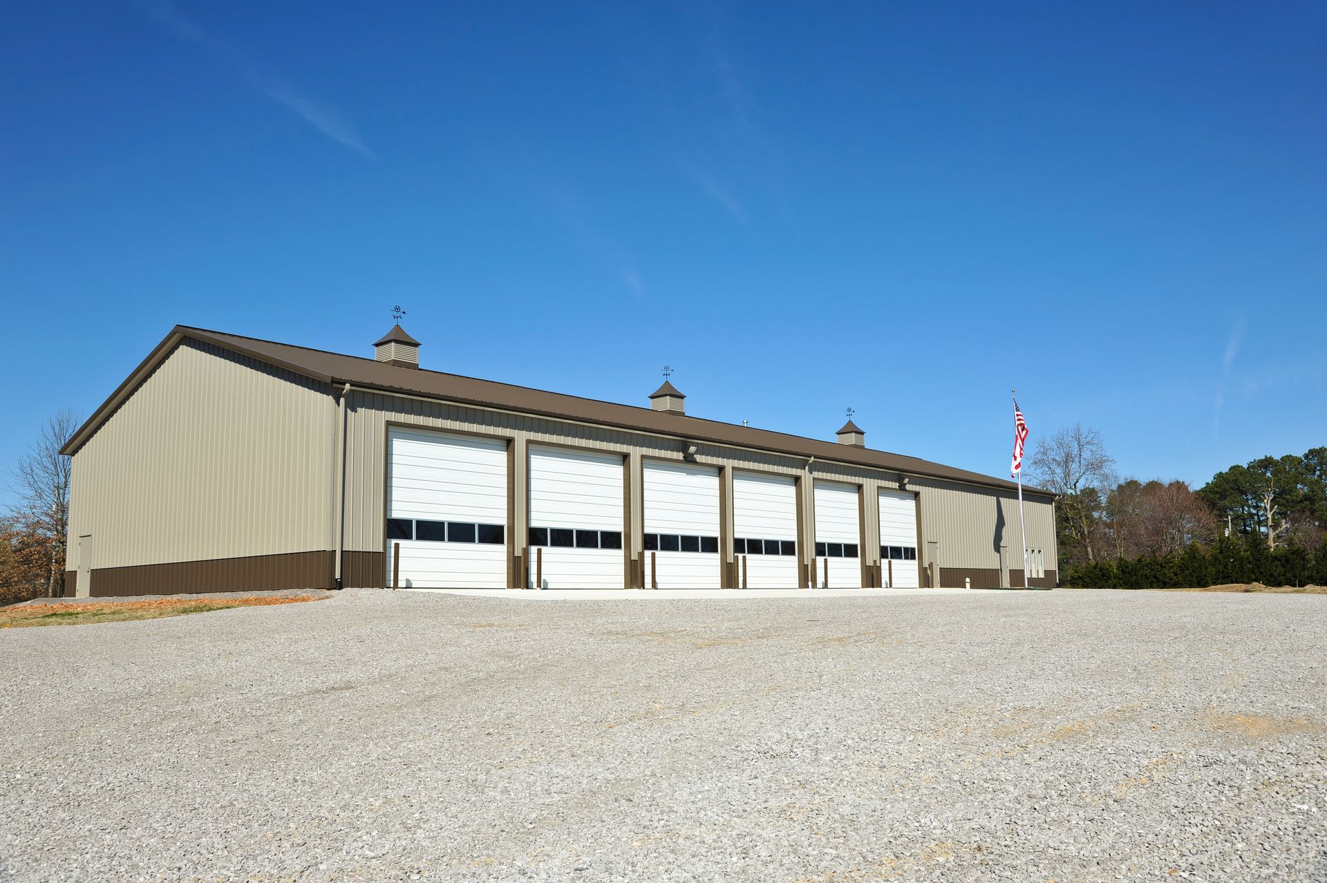 Tan metal building with six garage doors against a clear blue sky. A gravel lot is in the foreground.