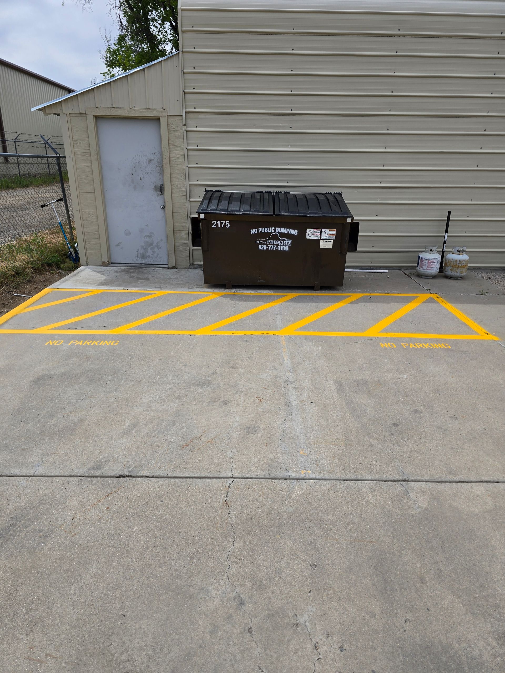 Dumpster beside a beige building in a yellow striped no-parking zone on concrete pavement