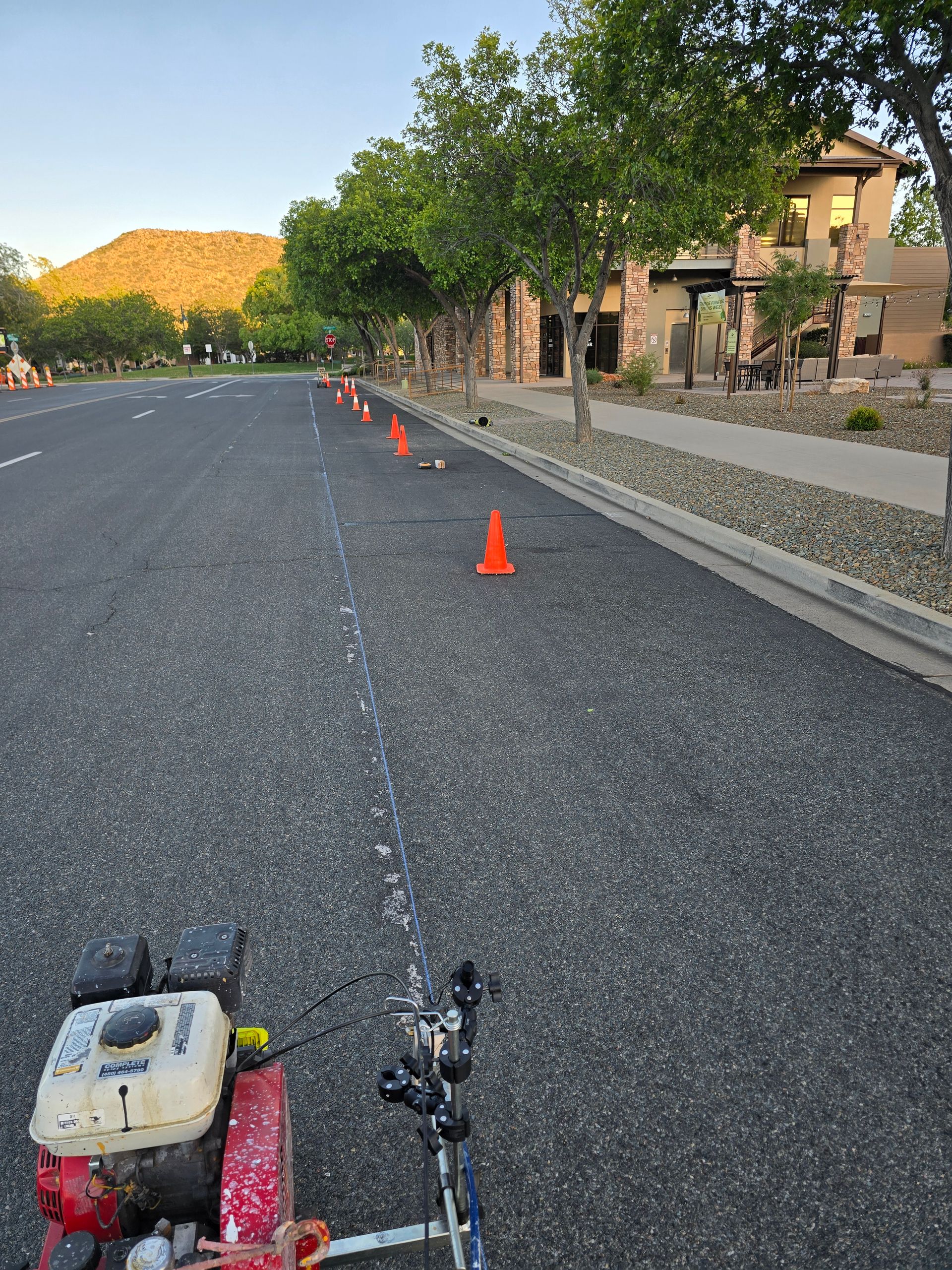 Roadway closed with orange cones lining the street beside a sidewalk and houses at sunset.