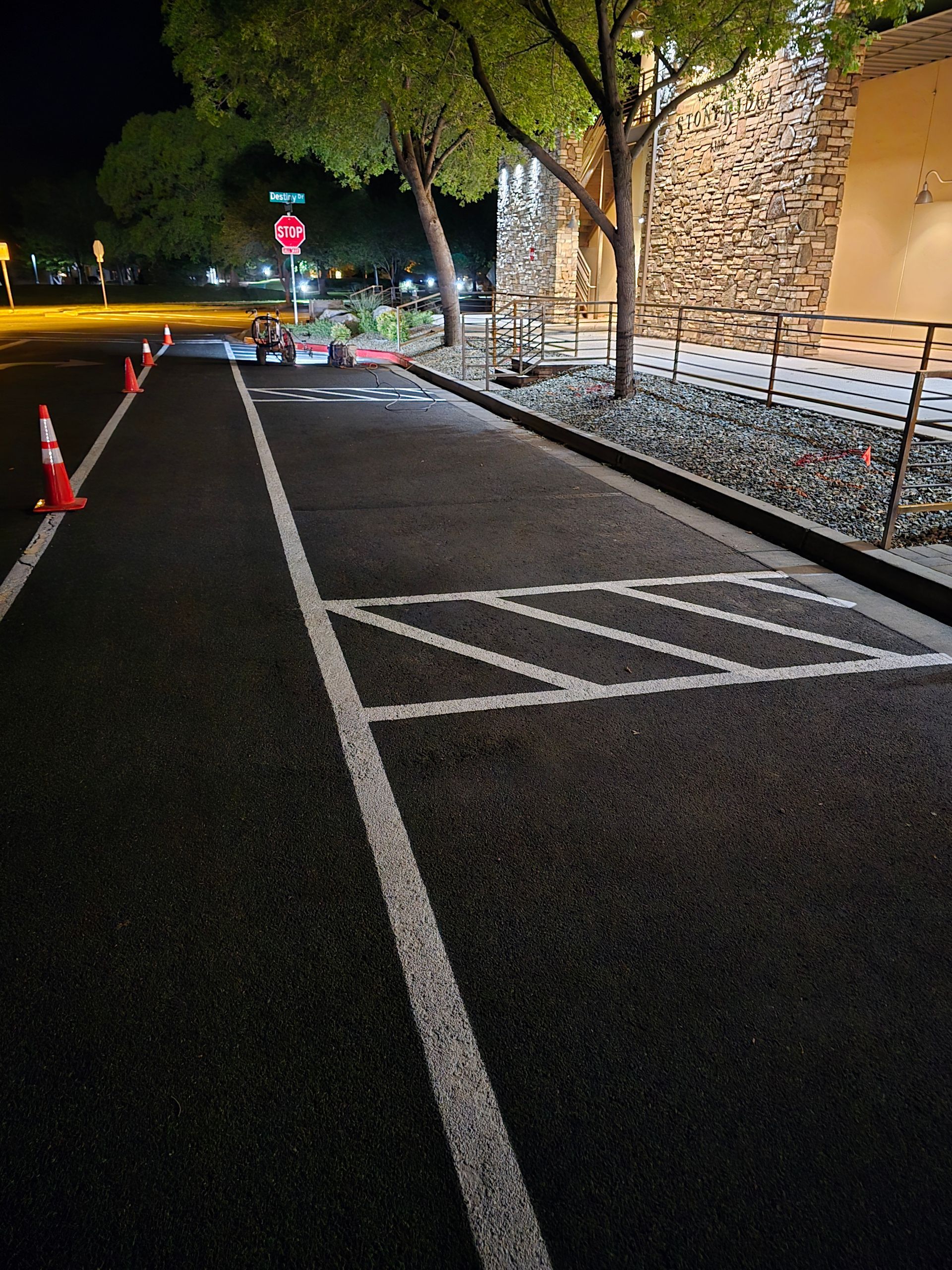Nighttime sidewalk beside a stone building, with road cones, a red stop sign, and marked pedestrian crossing lines.