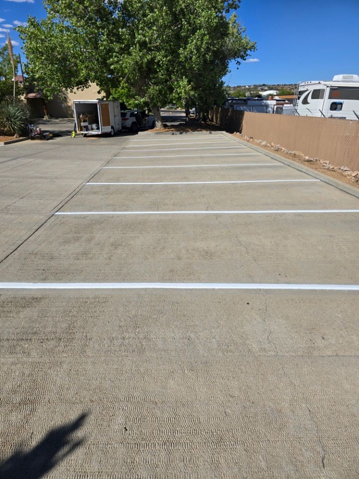 Empty paved parking lot with white striped spaces, trees, and RVs along a tan fence.