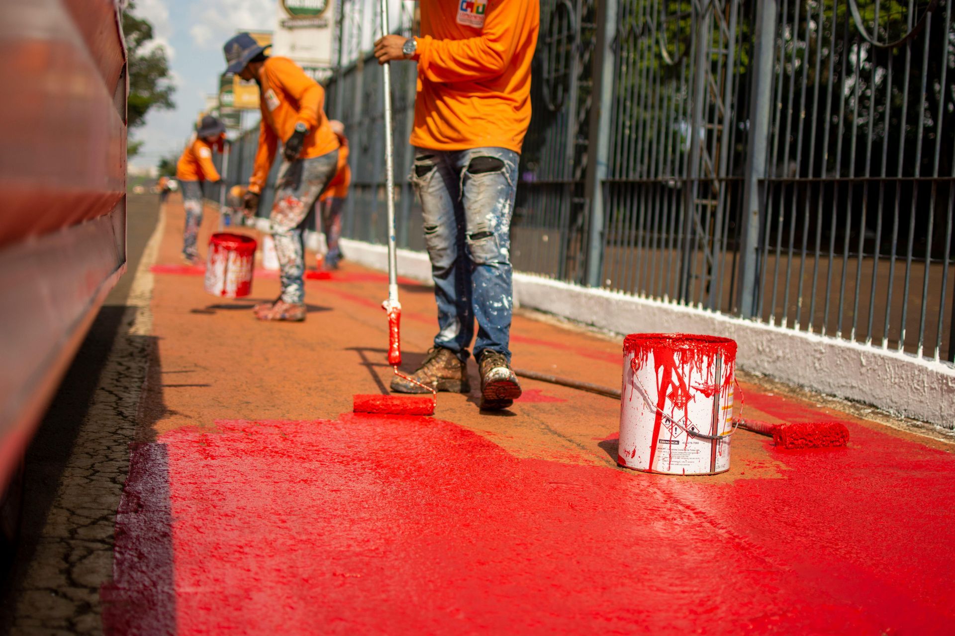 Workers painting a red pavement strip beside a fence, with paint buckets and rollers.