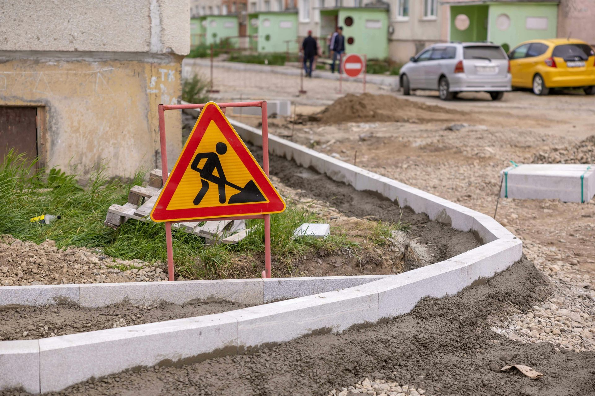 Construction warning sign beside a muddy road and curb under repair, with parked cars in the background