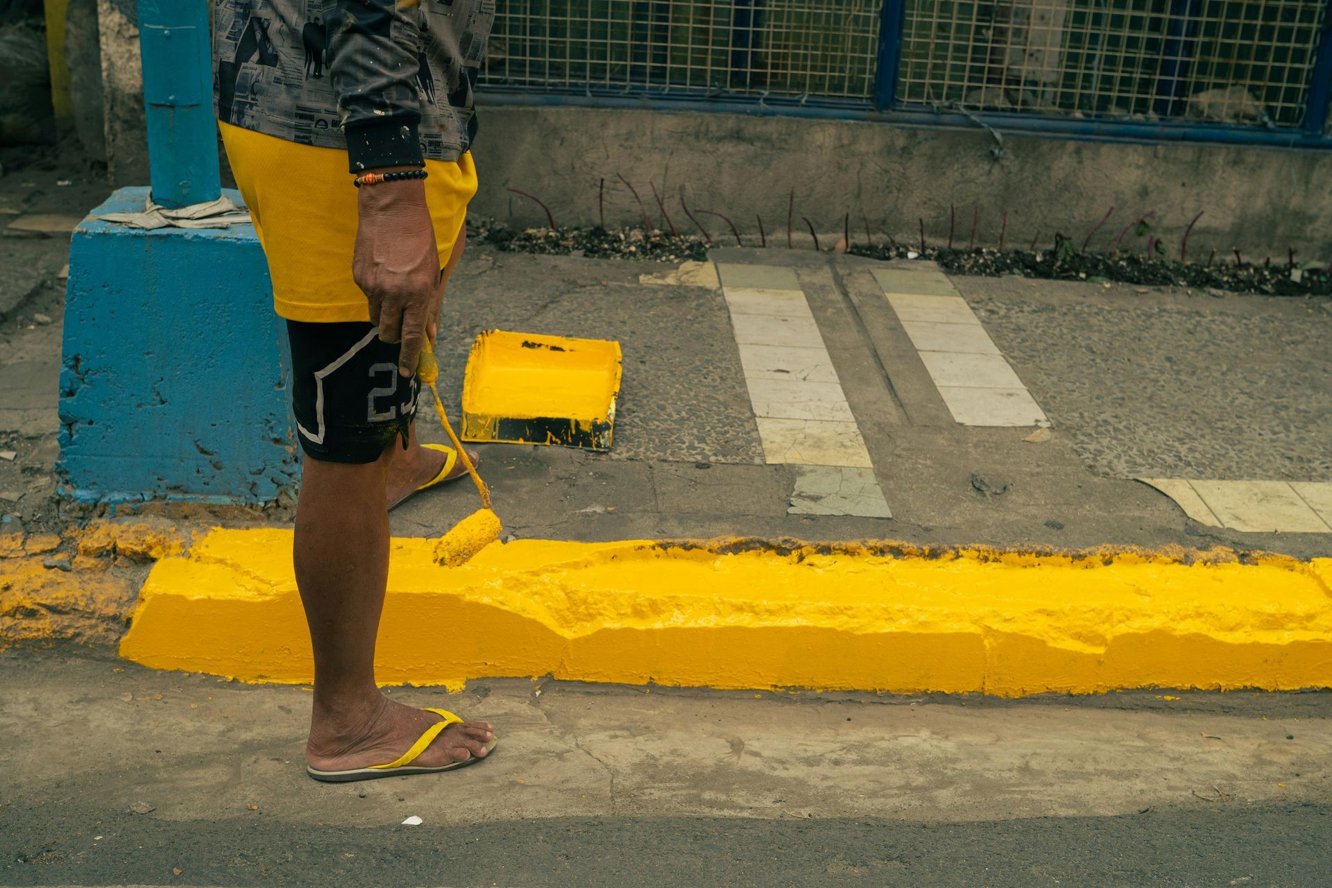 Person standing on a curb painted yellow, holding a cable near a blue wall and sidewalk.