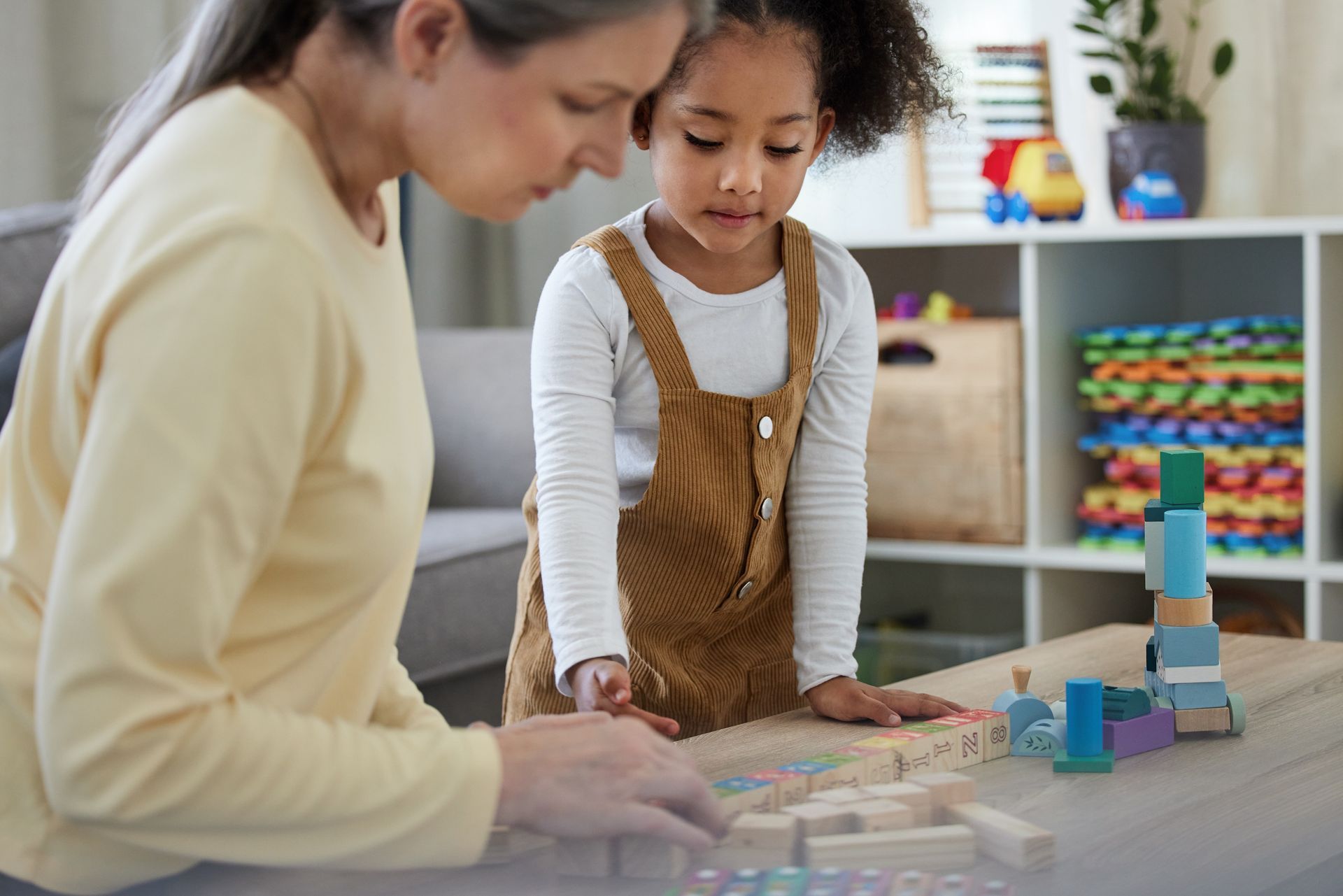 An adult and a child sitting at a table together, building a structure with wooden blocks in a room with toy shelves.