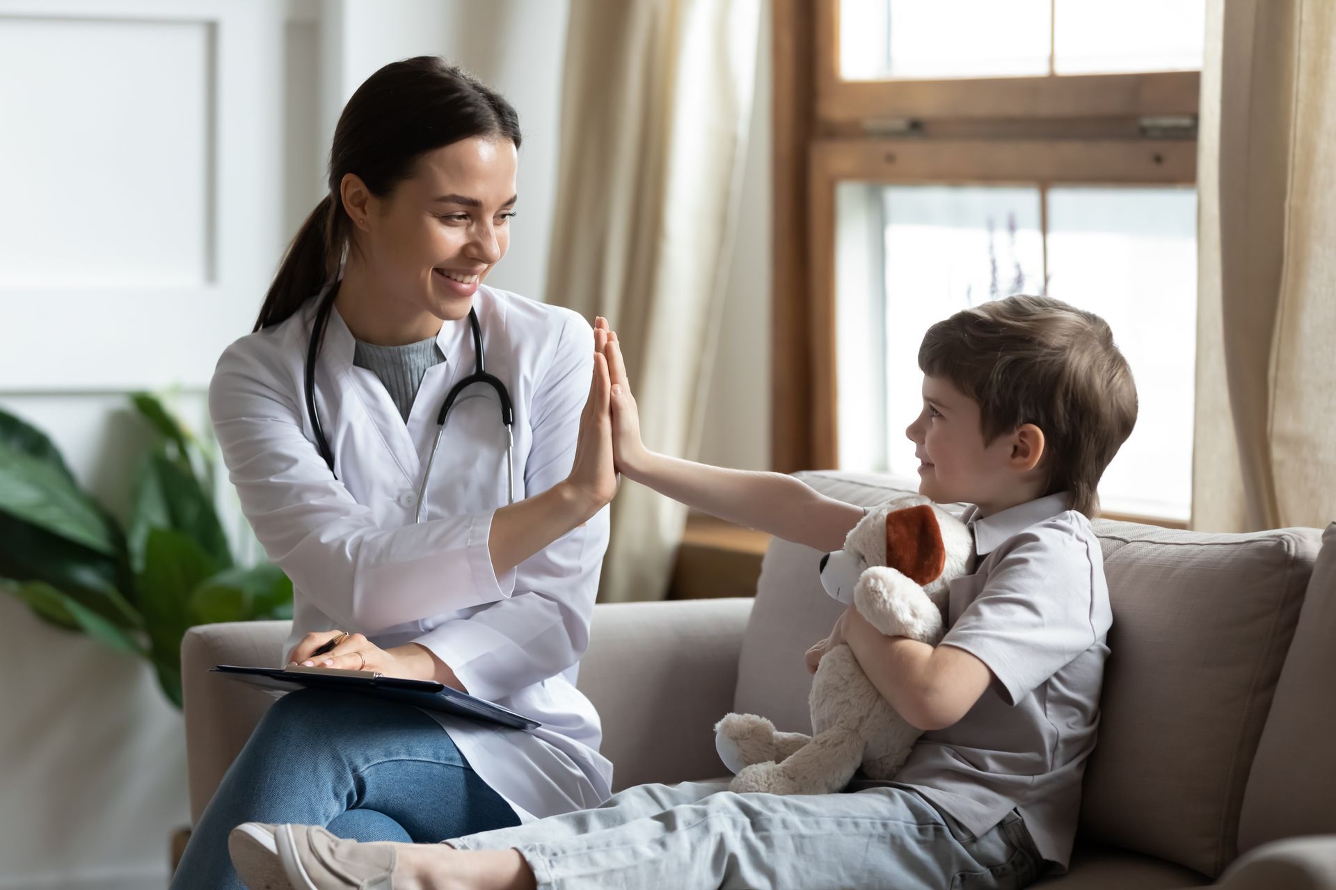 A clinician in a white coat gives a high-five to a young child holding a stuffed animal while sitting on a couch.