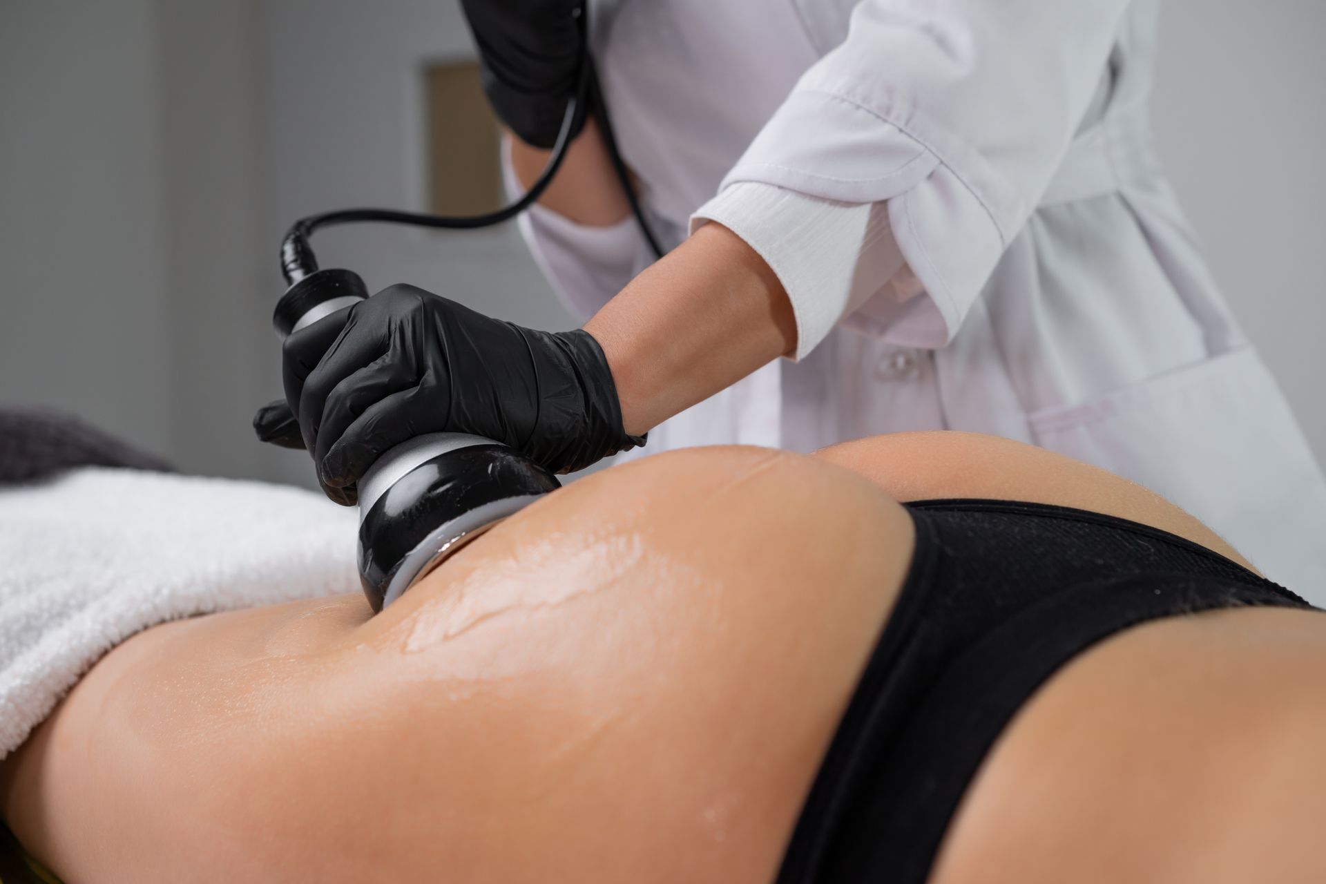 A practitioner in black gloves uses a cosmetic ultrasound device on a client's gluteal area in a clinic setting.
