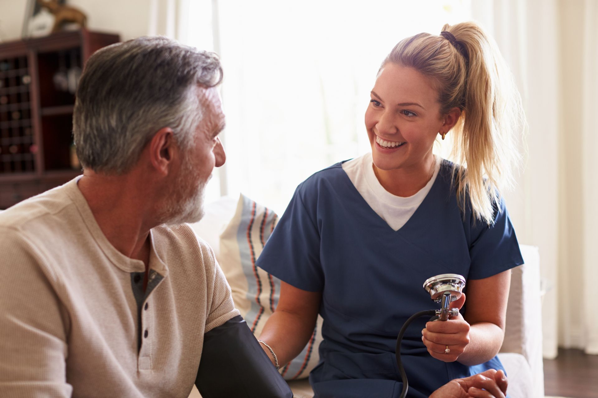 A smiling healthcare worker in blue scrubs checks a person’s blood pressure with a stethoscope in a home setting.
