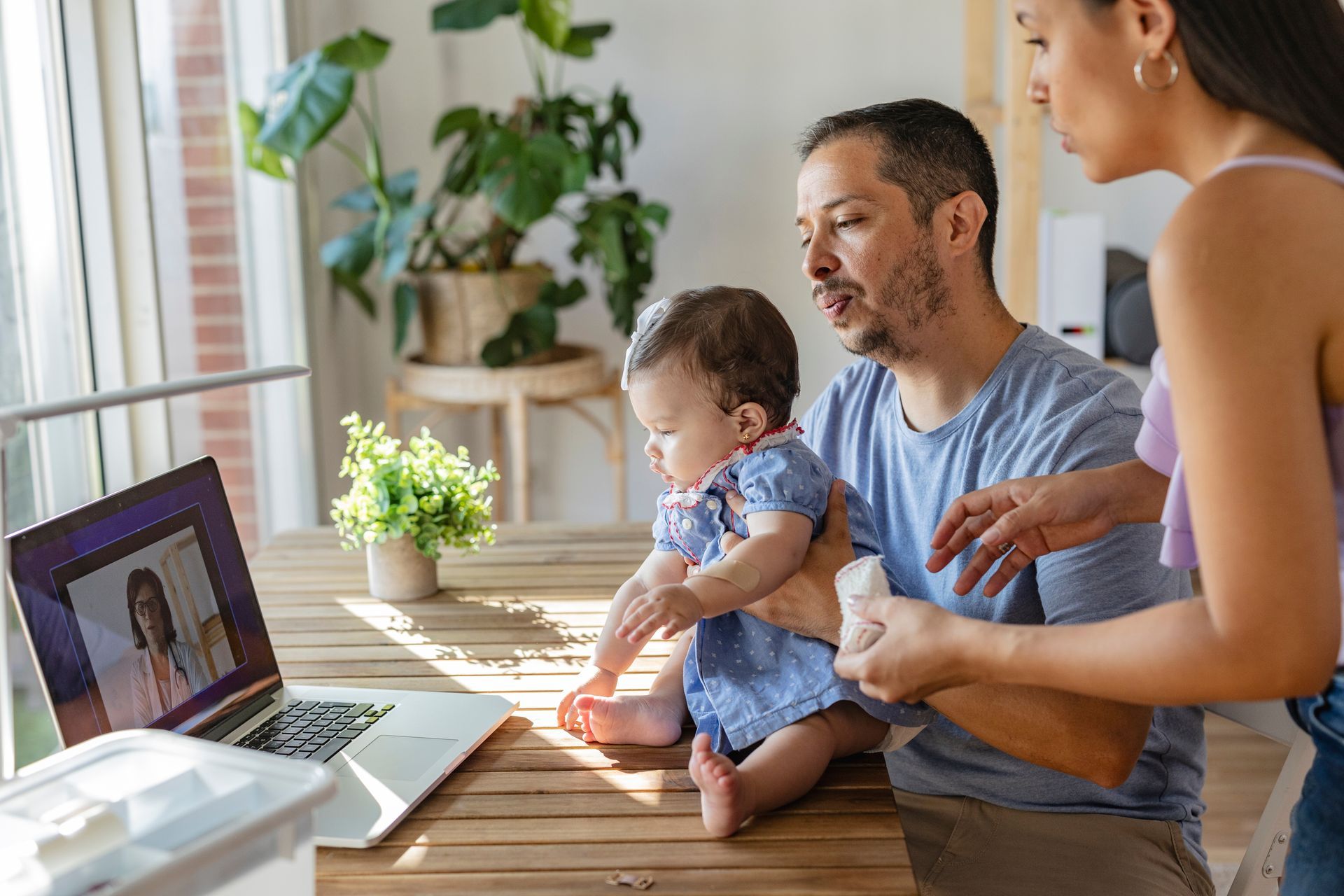 Parents with a baby on their lap looking at a video call on a laptop at a wooden table in a sunlit room.
