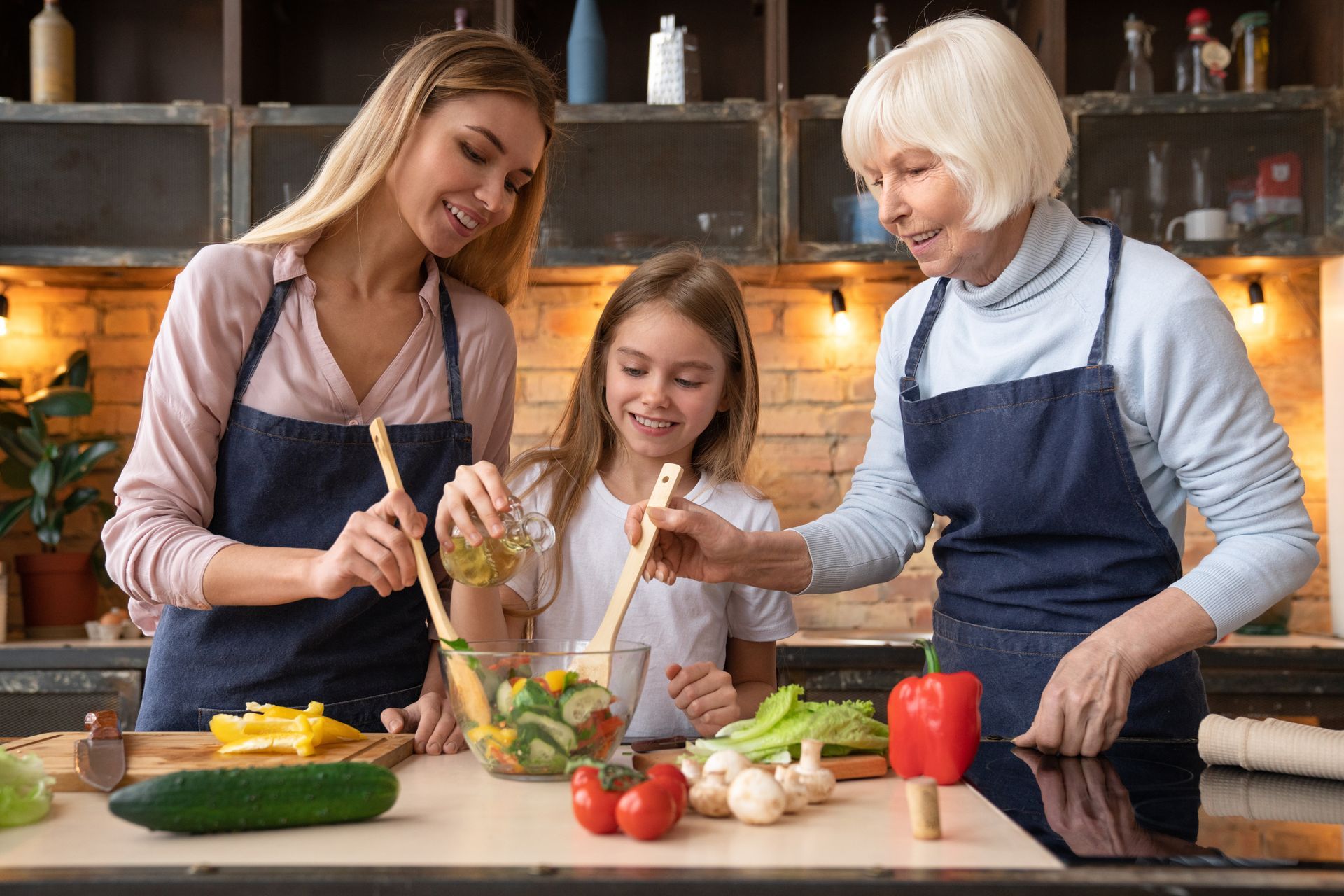 Three people wearing aprons prepare a salad together in a kitchen, smiling as they mix vegetables in a glass bowl.