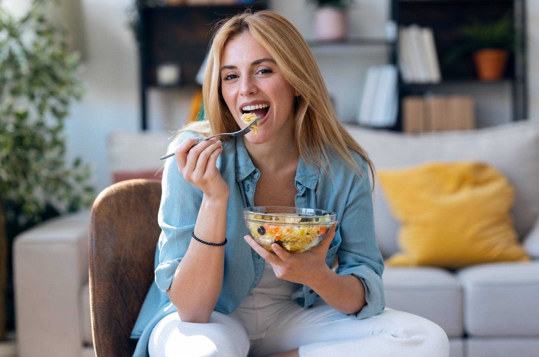 Woman smiling and eating salad on a couch in a bright living room