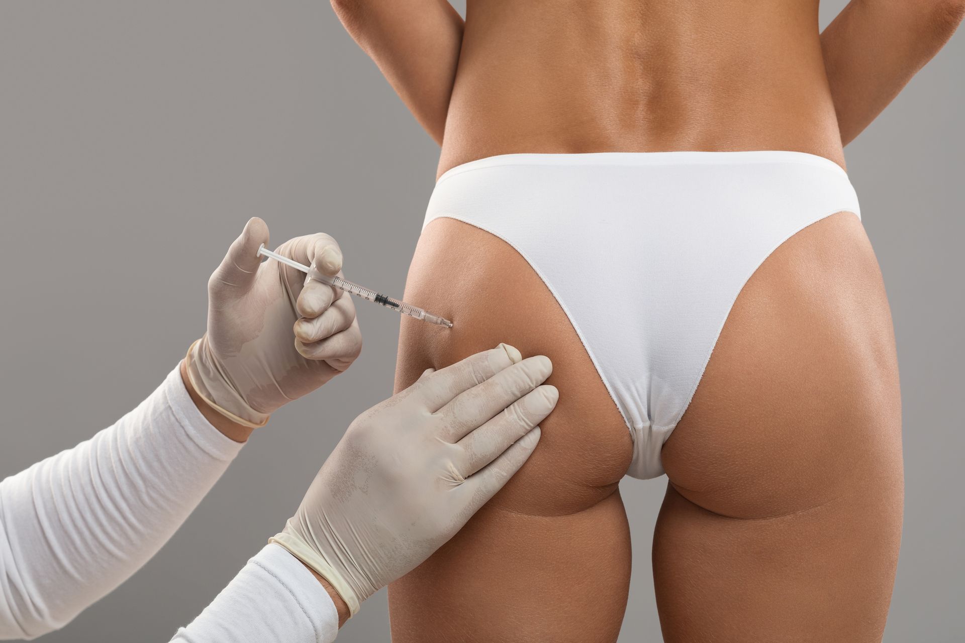 Gloved hands injecting a white syringe into a person’s buttock against a gray background