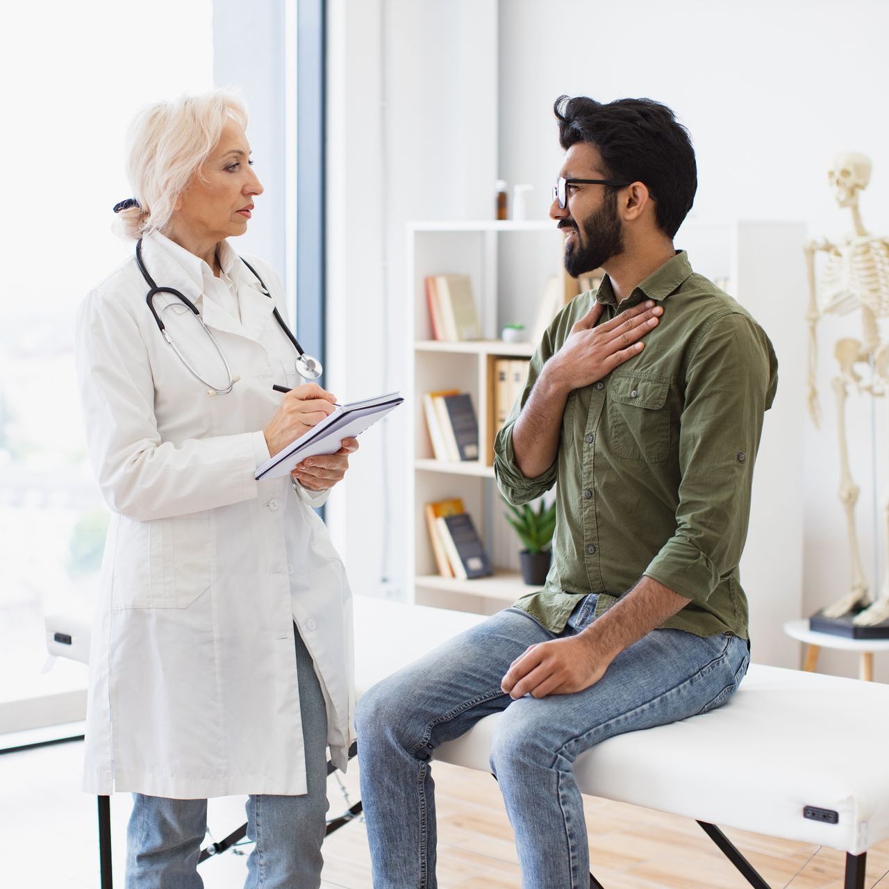 Doctor speaking with a seated patient in a bright clinic, holding a clipboard and touching his chest.