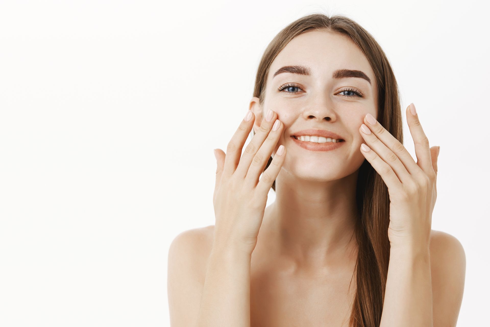 Smiling woman gently applying skincare to her cheeks against a white background