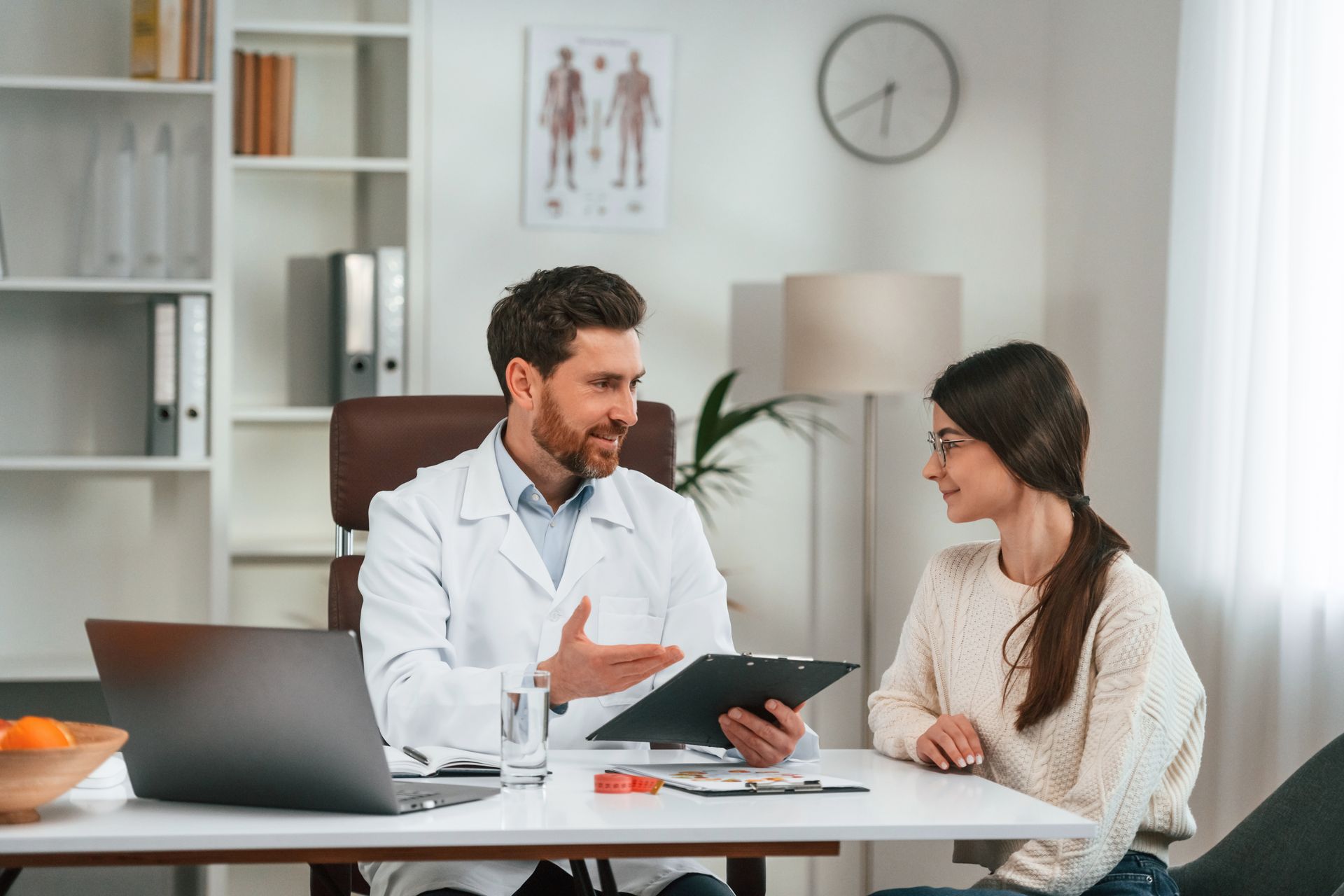 Doctor consulting with a woman at a desk in a bright office, holding a tablet.