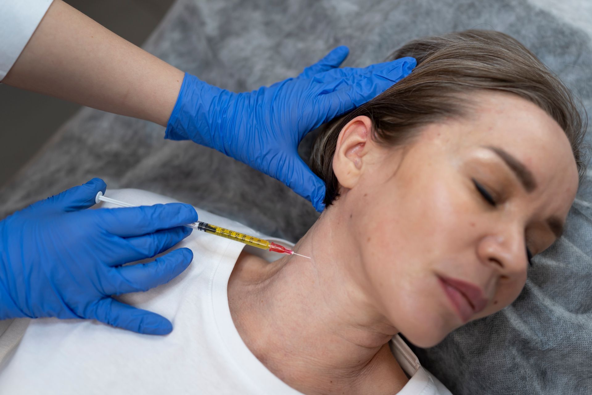 Gloved hands perform a neck injection on a reclining patient in a clinical setting