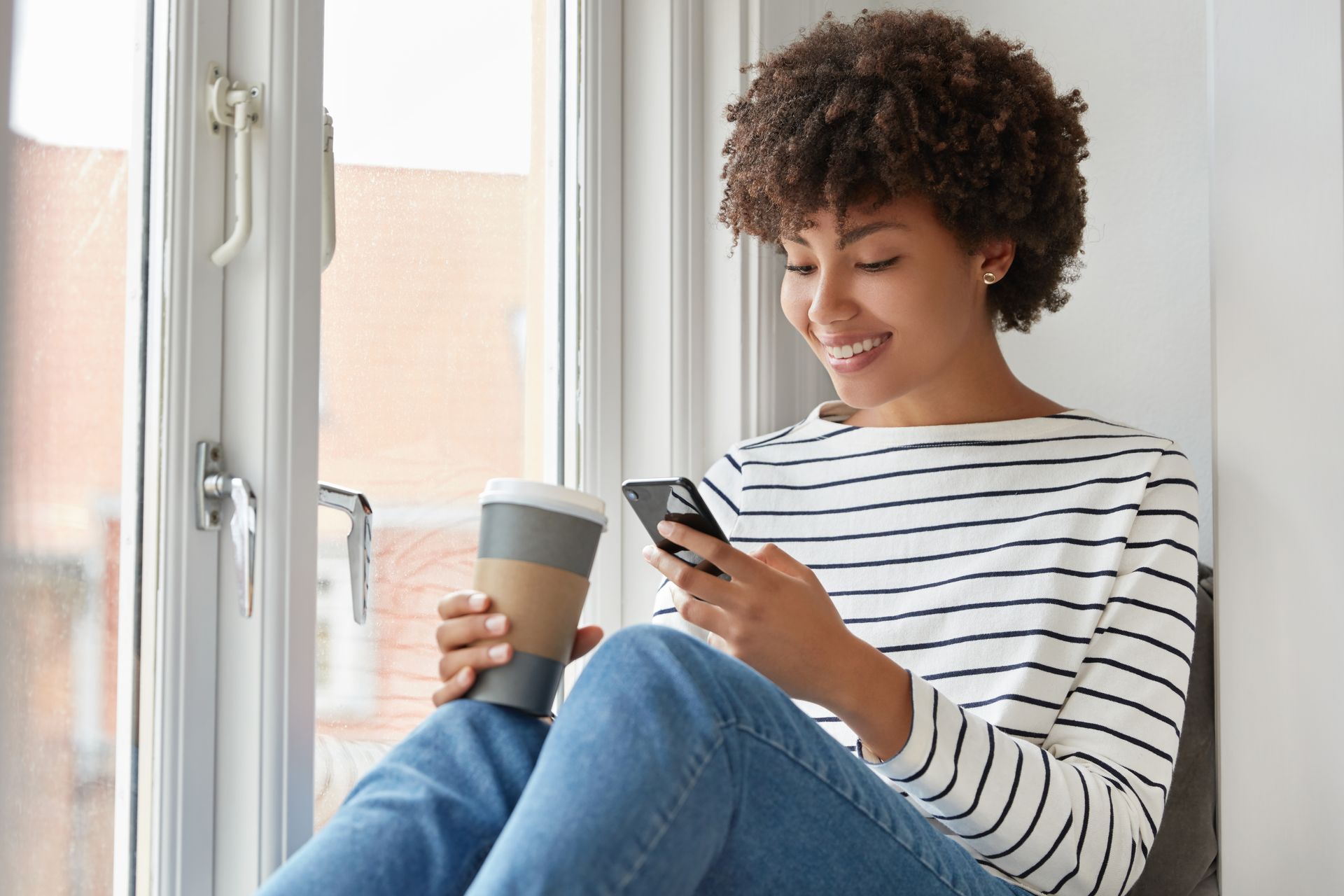 Smiling person in a striped shirt sitting by a window, holding a coffee cup and looking at a phone