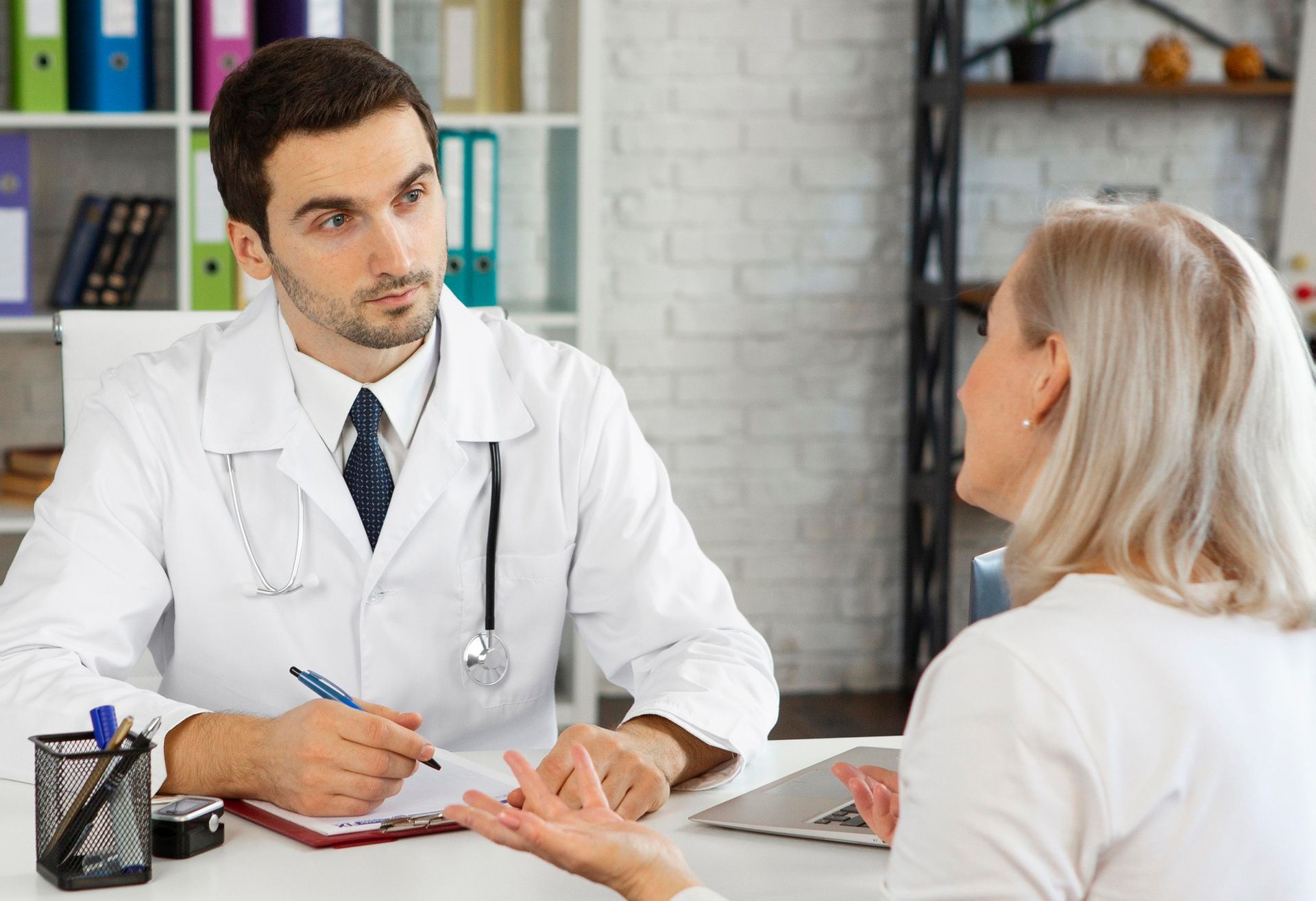 Doctor talking with a patient in a clinic office, both seated across a desk.