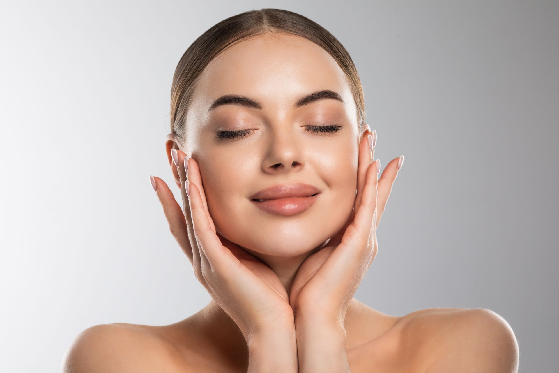 Smiling woman with closed eyes gently touching her face against a light background