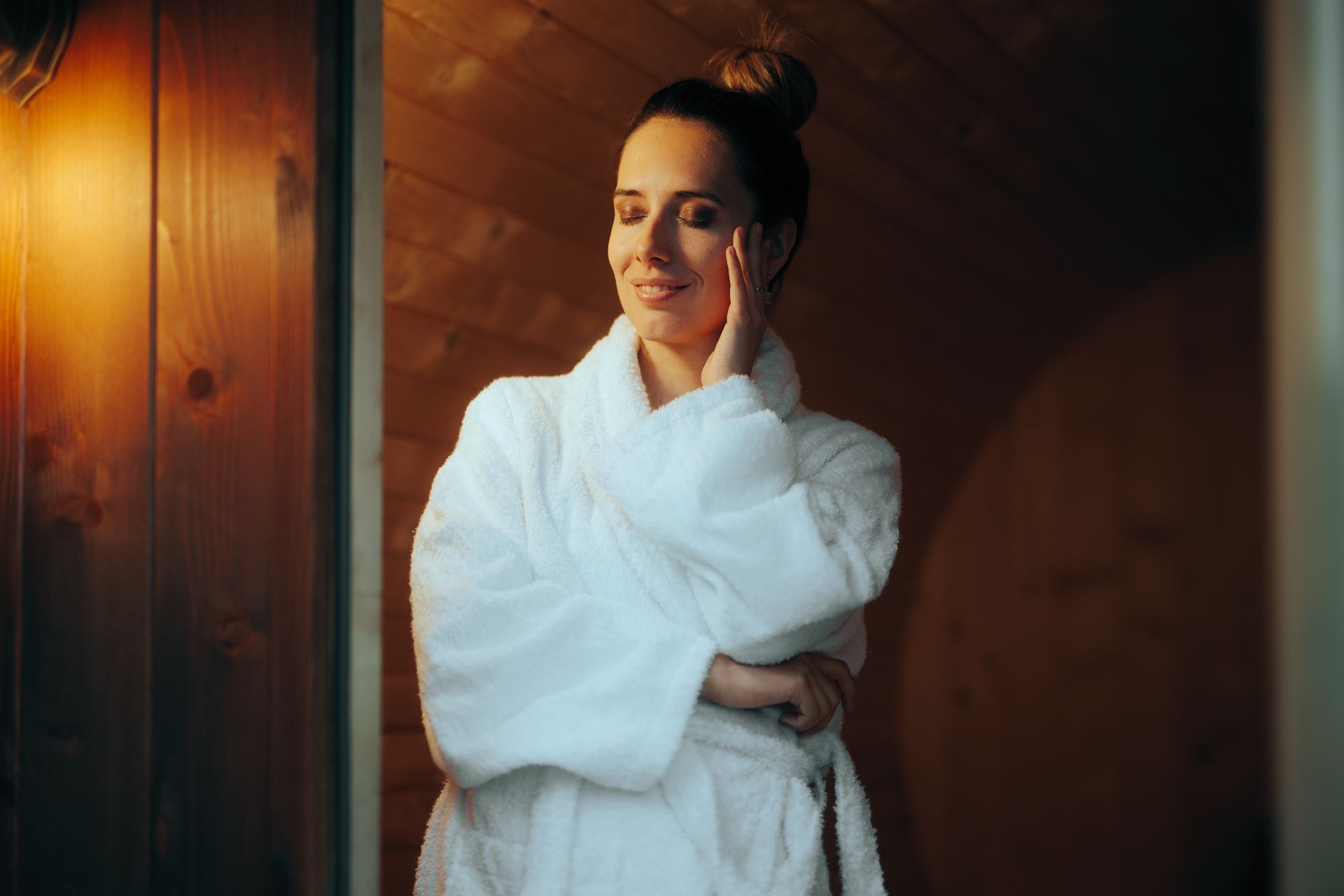 Person in a white robe smiling in a wooden spa room with warm lighting.