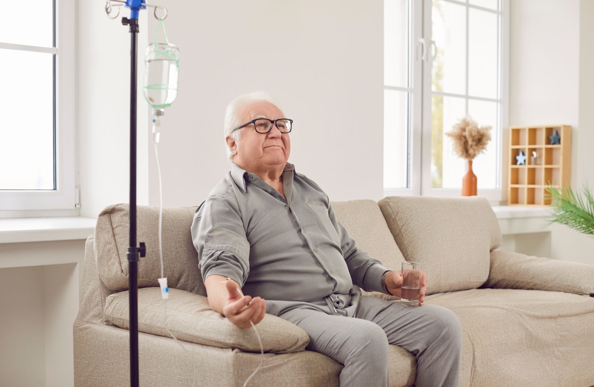 Elderly man seated on a beige sofa beside an IV stand in a bright living room, resting with a calm expression