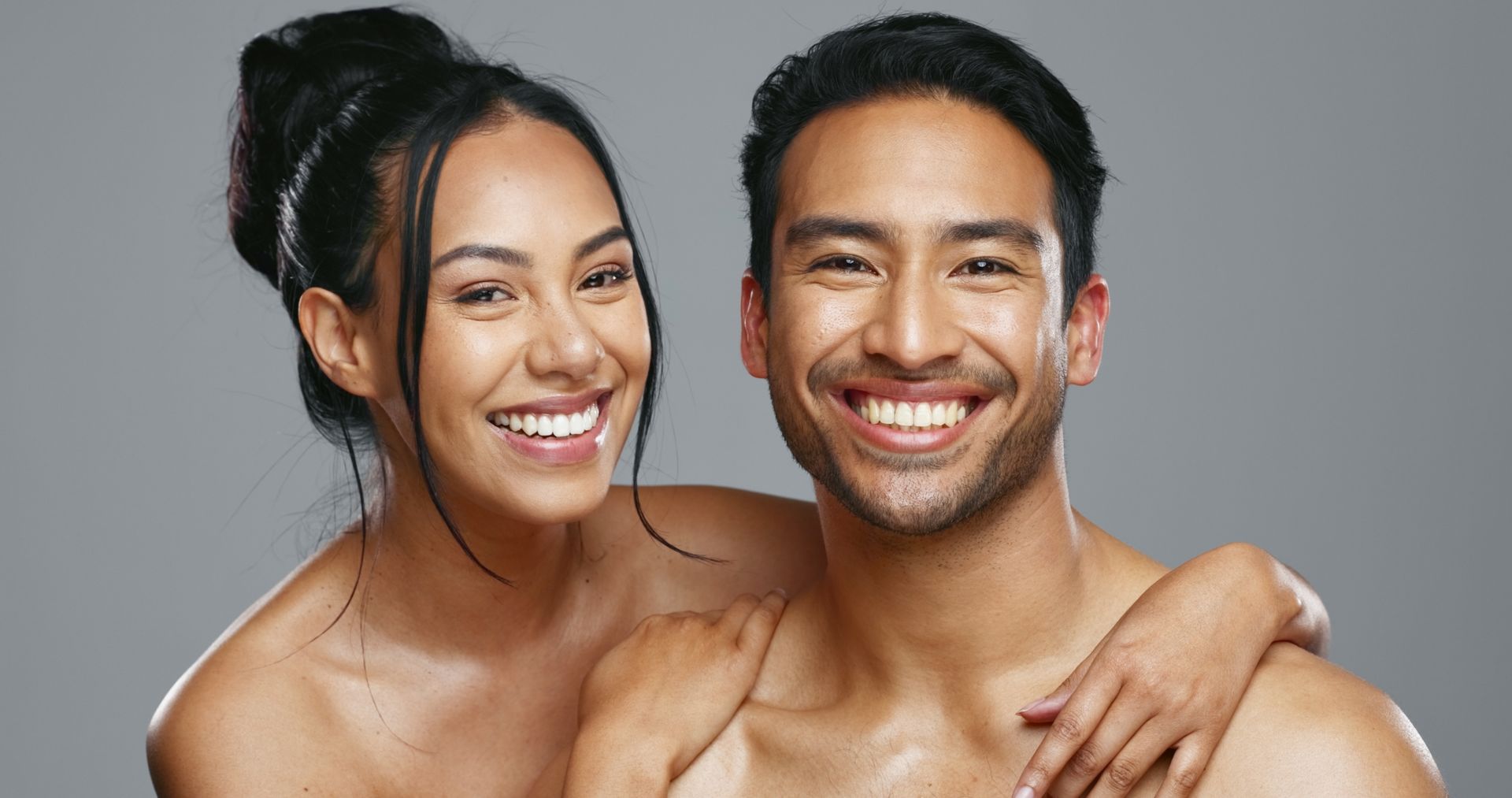 Smiling shirtless couple posing against a gray background, woman’s arm around man’s shoulders