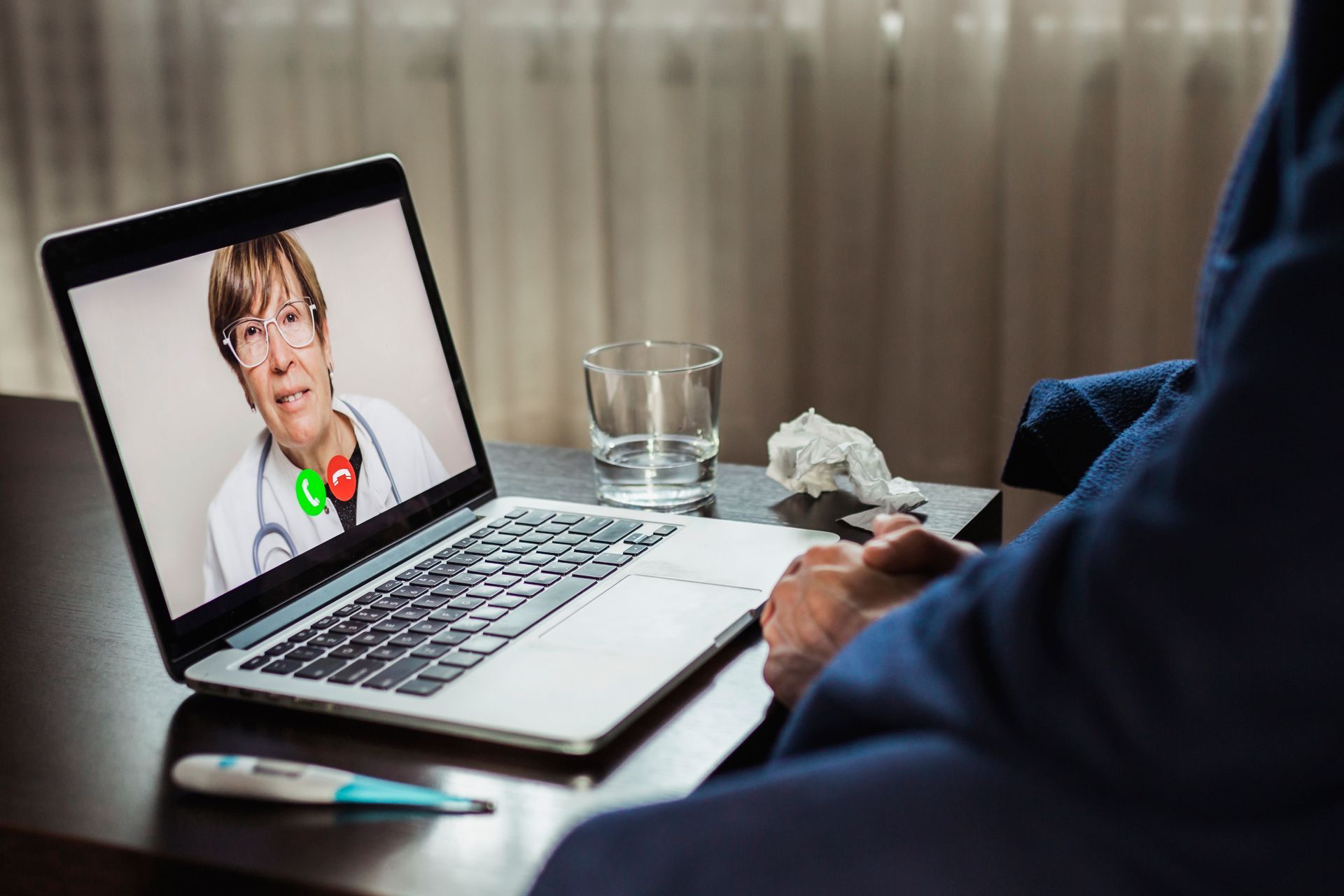 Laptop showing a video call with a doctor, with a glass of water and a tissue box on the desk.